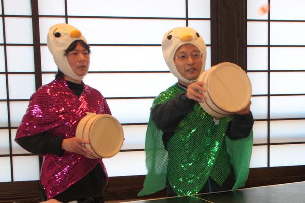 アヒルのような被り物をして緑と赤の衣装を身にまとった男性が桶を持って構えている写真